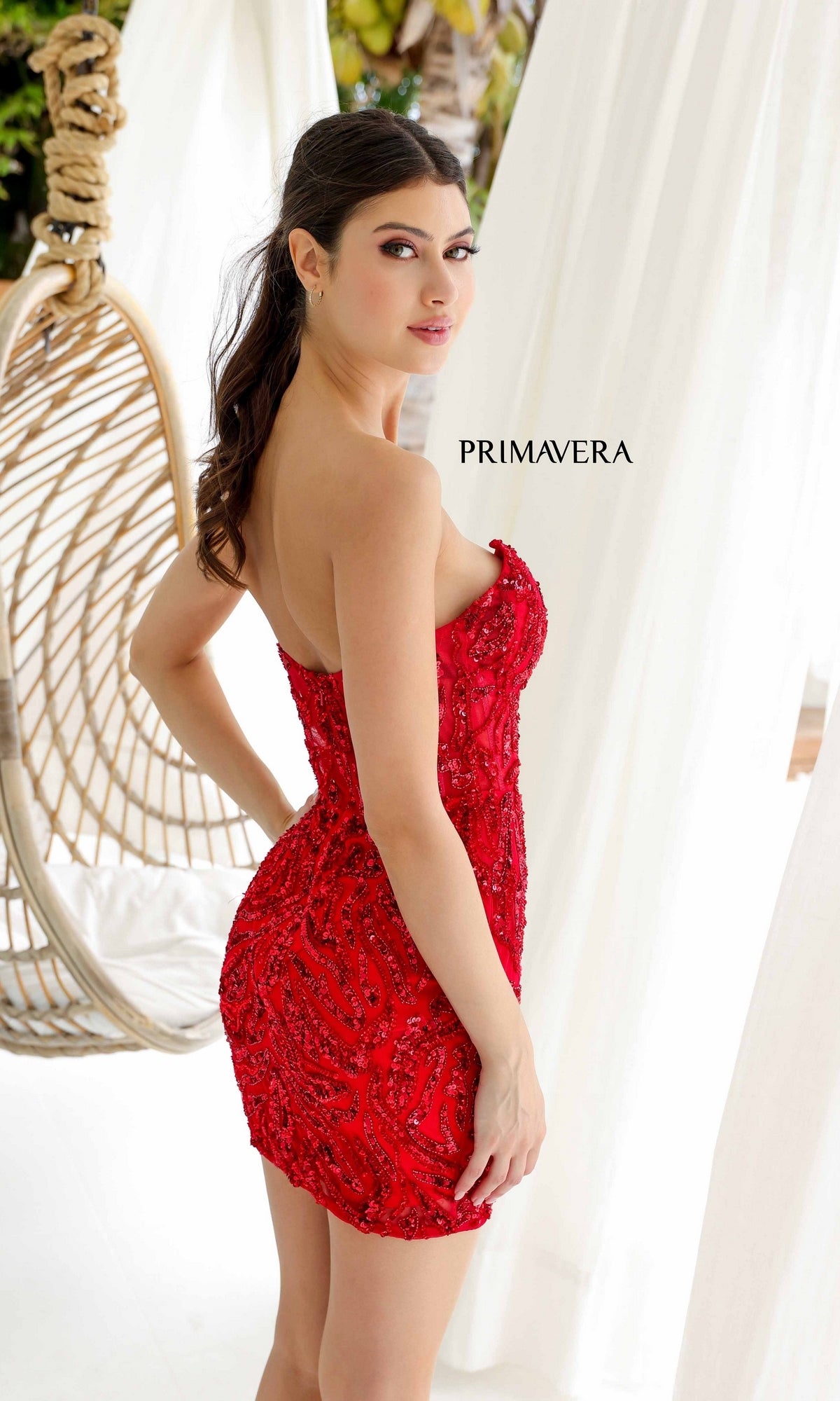 A woman models the Short Homecoming Dress 4222 by Primavera, featuring a strapless beaded mini dress design, as she stands beside a hanging chair and looks over her shoulder with